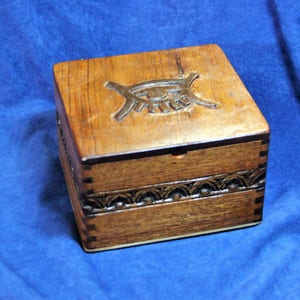 May include: A wooden box with a carved eye design on the lid. The box has a dark brown finish and intricate carvings around the edges.