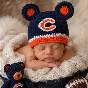 Op de afbeelding: Een marineblauwe en oranje gehaakte muts met berenoren en een wit letter C-logo, vergezeld door een bijpassende gehaakte teddybeer met een C-logo en een bruin-witte gehaakte football met witte stiksels.