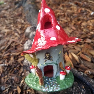 May include: A whimsical fairy house made of clay, painted red with white polka dots, and shaped like a mushroom. The house has a small door and a window, and a skull charm hangs above the door. The house is sitting on a bed of green moss.