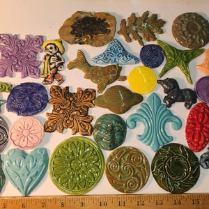 May include: A collection of various ceramic shapes and figures in diverse colors and textures, including floral, animal, and abstract designs, arranged on a surface next to a wooden ruler measuring up to 18 inches in length.