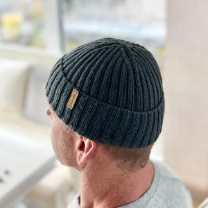 Op de afbeelding: Donkergrijze geribbelde gebreide beanie met een klein bruin leren label. De hoed is een klassieke stijl, geschikt voor koud weer. Het label heeft de tekst "Woolster".