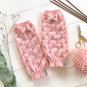 May include: A pair of pink knitted leg warmers with a delicate lace pattern and a small bow at the top. The leg warmers have a white tag with the word "Wool Addict" on them.