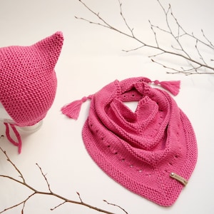 May include: A pink knitted hat with a pointed top and a matching pink knitted scarf with tassels. The scarf has a small leather tag with the word "Wool & The" printed on it.