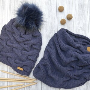 May include: A dark blue knitted hat with a faux fur pom pom and a matching dark blue knitted infinity scarf. The hat and scarf are laid out on a light wood surface with wooden knitting needles.