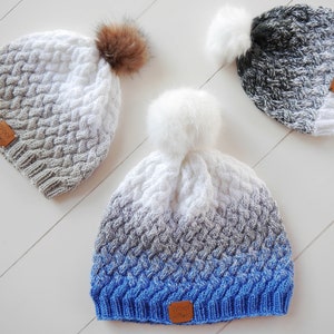 Puede incluir: Tres gorros de punto con pompones. Los gorros son degradados, con un degradado de gris a blanco, un degradado de negro a blanco y un degradado de azul a blanco. Cada gorro tiene una pequeña etiqueta de cuero marrón con la palabra "Wool" en ella.