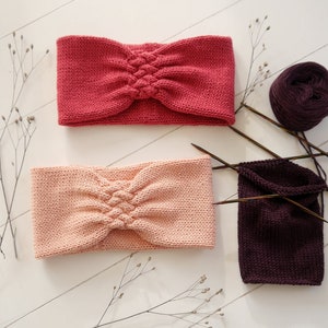 May include: Two knitted headbands in shades of pink and red are displayed. The headbands have a braided knot design in the center. A ball of dark purple yarn, knitting needles, and a partially knitted piece are also visible, suggesting a handmade craft project.