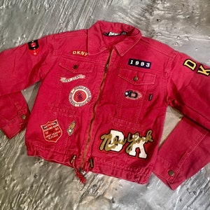 Puede incluir: Chaqueta vaquera roja con varios parches, incluyendo "DKNY Jeans", "National Bowling Assn" y "New York DK". La chaqueta tiene una cremallera y un cordón en la parte inferior.