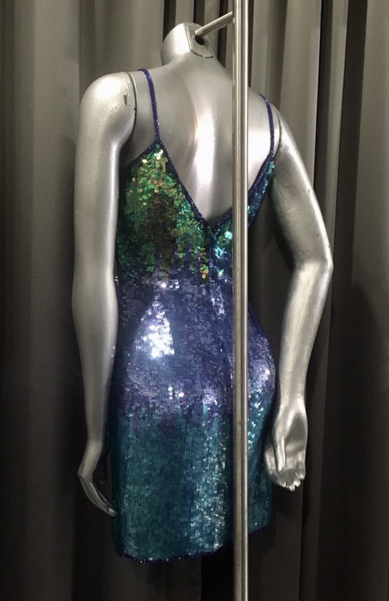 Może przedstawiać: Kr&oacute;tka, bezrękawowa sukienka z cekinami w kolorze zielonym, niebieskim i fioletowym. Sukienka ma dekolt w kształcie litery V i dopasowany kr&oacute;j.