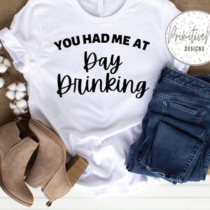 K&ouml;nnte beinhalten: Wei&szlig;es T-Shirt mit dem Text "You had me at Day Drinking" in schwarzer Schrift. Das Shirt liegt flach auf einer wei&szlig;en Oberfl&auml;che mit einem Paar braunen Wildlederstiefeln und einer blauen Jeans.