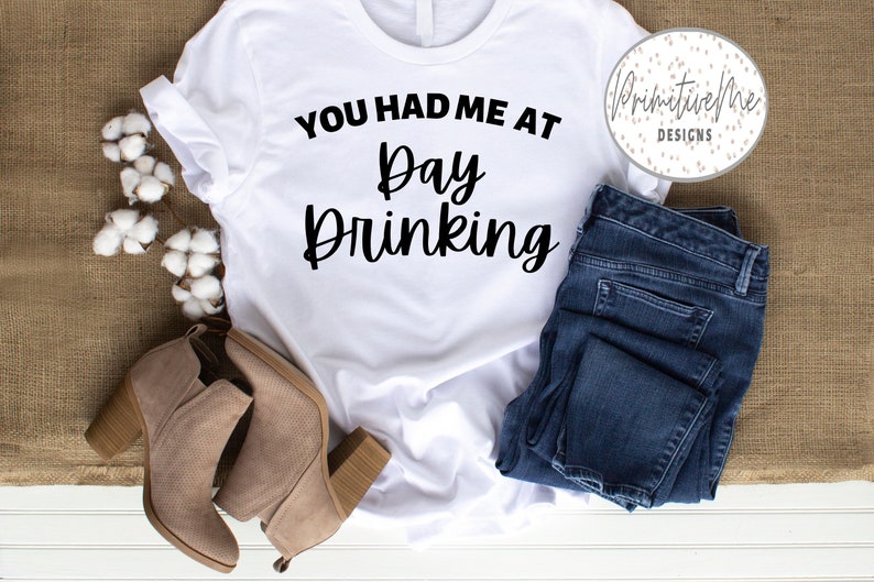 K&ouml;nnte beinhalten: Wei&szlig;es T-Shirt mit dem Text "You had me at Day Drinking" in schwarzer Schrift. Das Shirt liegt flach auf einer wei&szlig;en Oberfl&auml;che mit einem Paar braunen Wildlederstiefeln und einer blauen Jeans.