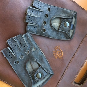 May include: Two pairs of black leather driving gloves with perforated detailing and a snap closure. The gloves are on a brown leather surface with the text "A.B.R.A.M." in a brown swirl design.