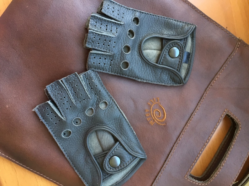 May include: Two pairs of black leather driving gloves with perforated detailing and a snap closure. The gloves are on a brown leather surface with the text "A.B.R.A.M." in a brown swirl design.