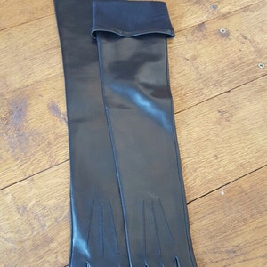 May include: A pair of long, black leather gloves, resting on a wooden surface. The gloves extend well past the elbow, with a black elastic band at the upper arm. The fingers are precisely stitched.
