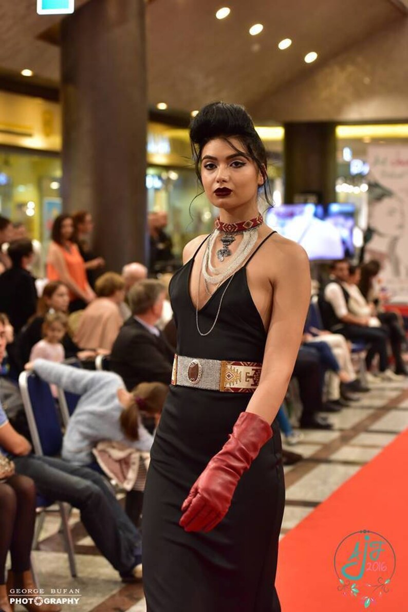 May include: A woman wearing a black dress with a red and silver belt, red gloves, and multiple silver necklaces. She is wearing red lipstick and has her hair styled in a high bun.