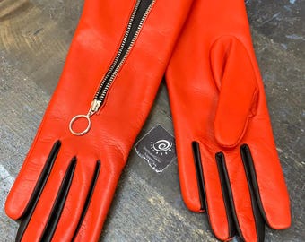 May include: A pair of bright orange leather gloves featuring black leather finger gussets and a prominent vertical silver-toned zipper closure on the back of the left glove, complete with a circular pull tab and a small square brand tag.