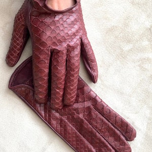 May include: A pair of burgundy leather driving gloves with a snakeskin pattern. The gloves are laid out flat on a white surface.