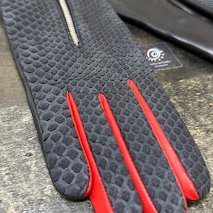 May include: Black leather gloves with a textured, scale-like pattern. The gloves have red accents along the fingers and a silver zip. A pair of scissors is visible in the background.