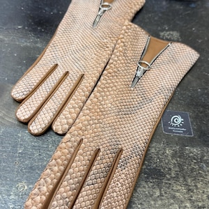 Puede incluir: Un par de guantes de cuero color canela con un patrón de piel de serpiente. Los guantes tienen una cremallera en la muñeca y ribetes marrones. Se muestran sobre una superficie oscura con retales de cuero y tijeras.