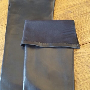 May include: Two black leather sleeves, one folded over, are displayed on a wooden surface. The folded sleeve has a black fabric cuff. The leather appears smooth and the lighting creates highlights on the surface.