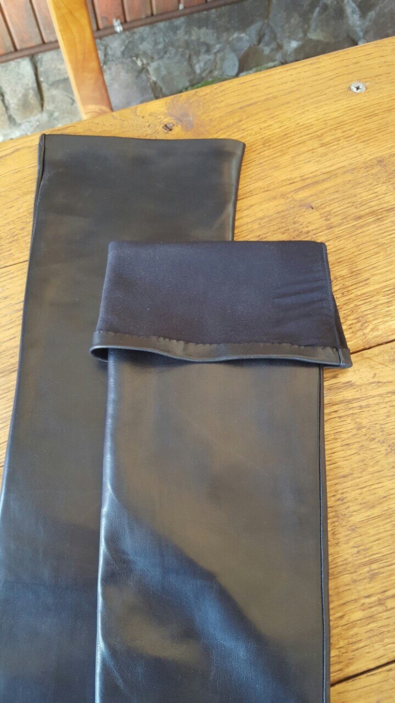 May include: Two black leather sleeves, one folded over, are displayed on a wooden surface. The folded sleeve has a black fabric cuff. The leather appears smooth and the lighting creates highlights on the surface.