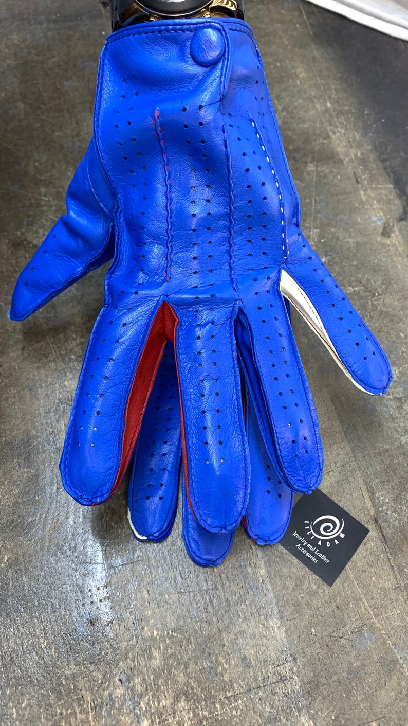 Pu&ograve; includere: Guanti da guida in pelle blu con cuciture rosse e dettagli bianchi. I guanti hanno un design traforato e un bottone al polso.