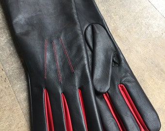 May include: Black leather gloves with red detailing on the fingers and red stitching. The gloves are long, extending past the wrist. The gloves are made of a smooth leather material.