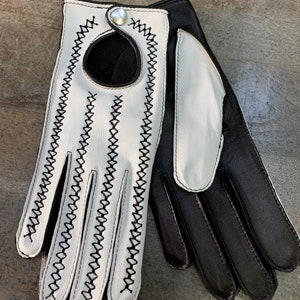 Puede incluir: Un par de guantes de conducir de cuero en blanco y negro. Los guantes tienen un patrón de costura decorativo y un botón en la muñeca.