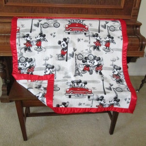 May include: A white and grey fleece blanket with a red satin border. The blanket features a repeating pattern of Mickey Mouse and Minnie Mouse in a variety of poses, including holding hands, riding a bicycle, and driving a red car. The blanket is ideal for a child's room or for snuggling up on the sofa.