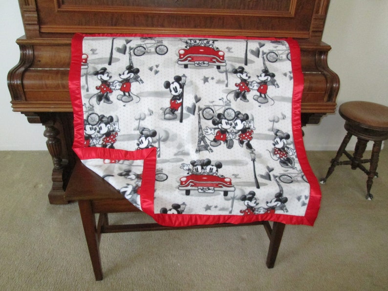 May include: A white and grey fleece blanket with a red satin border. The blanket features a repeating pattern of Mickey Mouse and Minnie Mouse in a variety of poses, including holding hands, riding a bicycle, and driving a red car. The blanket is ideal for a child's room or for snuggling up on the sofa.