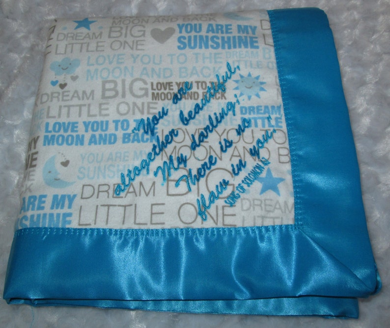 May include: A baby blanket with a white background and blue text that reads "You are my sunshine" and "Love you to the moon and back." The blanket has a blue satin border and embroidered text that says "You are beautiful, my darling, there is no flaw in you."