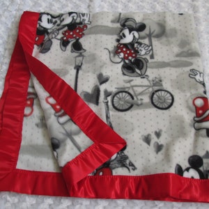 May include: White fleece blanket with a red satin border. The blanket features a repeating pattern of Mickey and Minnie Mouse in various poses, including riding a bicycle and holding hands. The background is a light grey with white polka dots and small hearts.