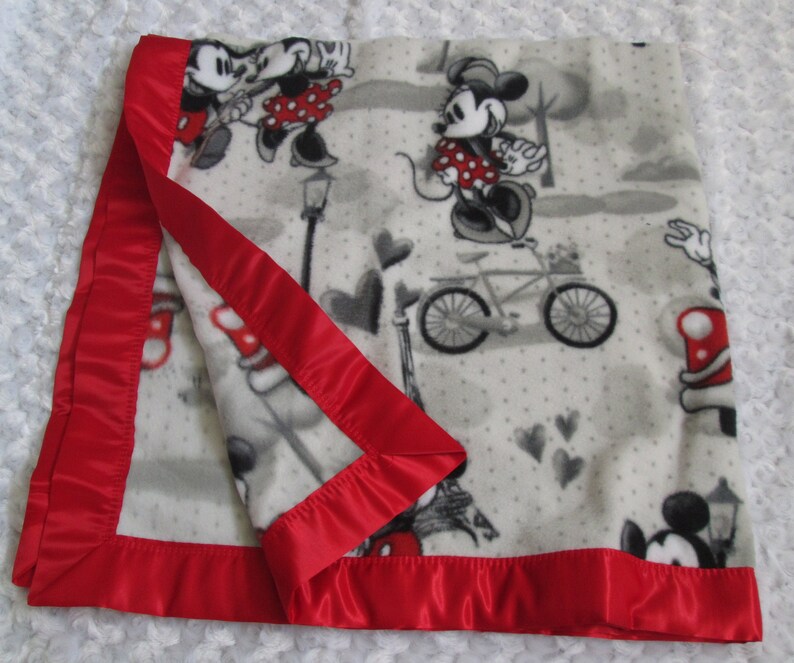 May include: White fleece blanket with a red satin border. The blanket features a repeating pattern of Mickey and Minnie Mouse in various poses, including riding a bicycle and holding hands. The background is a light grey with white polka dots and small hearts.