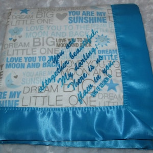 May include: A white baby blanket with a blue satin border. The blanket is covered in a repeating pattern of grey text that says "You are my sunshine", "Dream big little one", "Love you to the moon and back", and other similar phrases. The text is embroidered in a light blue colour. The blanket is folded in half and the border is visible on the bottom edge.