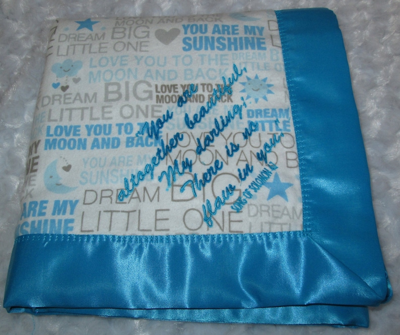 May include: A white baby blanket with a blue satin border. The blanket is covered in a repeating pattern of grey text that says "You are my sunshine", "Dream big little one", "Love you to the moon and back", and other similar phrases. The text is embroidered in a light blue colour. The blanket is folded in half and the border is visible on the bottom edge.