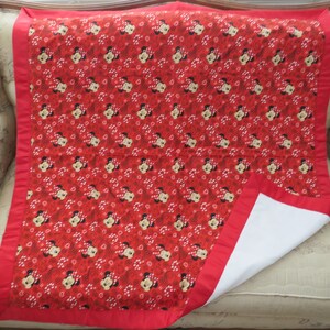 May include: A red and white baby blanket featuring a repeating pattern of Minnie Mouse faces and white polka dots. The blanket has a red border and a white underside. The blanket is folded over on a beige surface.