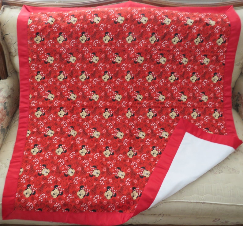 May include: A red and white baby blanket featuring a repeating pattern of Minnie Mouse faces and white polka dots. The blanket has a red border and a white underside. The blanket is folded over on a beige surface.