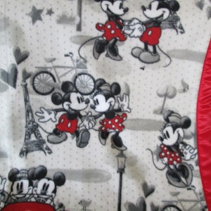 May include: A white fleece blanket with a red satin border features a repeating pattern of Mickey and Minnie Mouse in various poses. The pattern also includes hearts, stars, bicycles, and the Eiffel Tower.