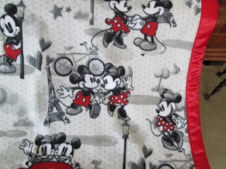 May include: A white fleece blanket with a red satin border features a repeating pattern of Mickey and Minnie Mouse in various poses. The pattern also includes hearts, stars, bicycles, and the Eiffel Tower.