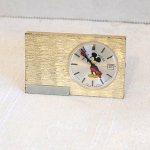 May include: A vintage gold-toned Mickey Mouse clock. The clock face features Mickey Mouse with red shorts and yellow shoes. The date is displayed as the number 27. The clock has a rectangular shape with a textured finish.