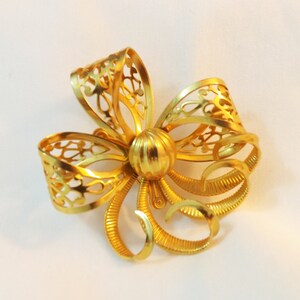 May include: A gold-toned metal brooch in the shape of a bow with intricate cut-out details. The bow has a round center with a textured surface.