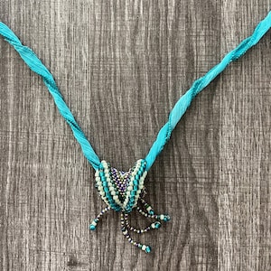 May include: A turquoise blue beaded necklace with a decorative knot at the center. The necklace is made of small, round beads in shades of turquoise, white, and black. The knot is a cluster of beads that are arranged in a spiral pattern.