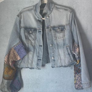 May include: Light blue denim jacket with patchwork sleeves. The jacket has a classic collar, button-down front, and two chest pockets. The sleeves are adorned with colorful, patterned fabric patches, adding a unique, artistic flair to the garment.