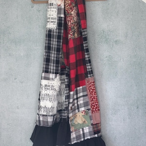 May include: A patchwork scarf with a variety of patterns, including red and black buffalo plaid, floral prints, and black and white checkered fabric. The scarf has a black ruffle at the bottom and is hanging on a wooden hanger.