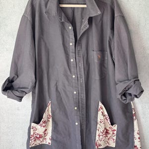 May include: Gray button-down shirt with floral pocket and hem accents. The shirt features a small embroidered logo on the chest and is displayed on a hanger. The sleeves are rolled up, and the pockets and hem have a cream and red floral pattern.