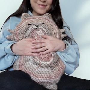 May include: A person holding a crocheted cat plush toy with brown, pink, and white stripes. The toy has a black embroidered face with a small X for eyes.