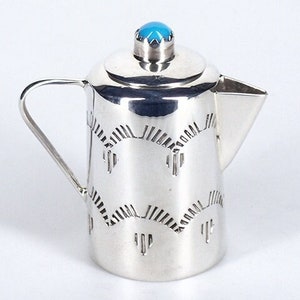 May include: A silver creamer with a turquoise stone on the lid. The creamer has a geometric design and a rounded body.