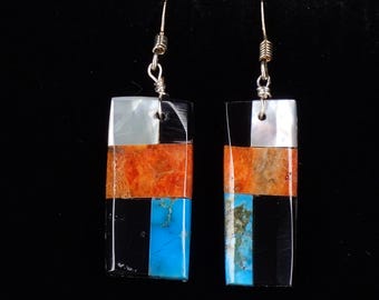 May include: A pair of rectangular drop earrings with silver hooks. Each earring has a layered design of white, orange, black, and turquoise-coloured stones. The earrings hang against a black background.