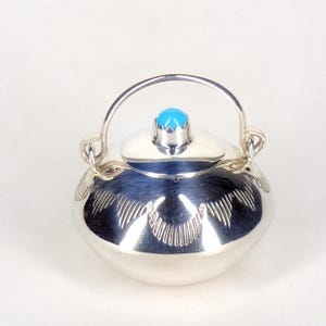 Op de afbeelding: Een kleine, zilverkleurige decoratieve pot met een handvat en deksel. Het deksel is bekroond met een kleine, ronde, turquoise gekleurde steen. De pot heeft geëtste ontwerpen rond de basis.