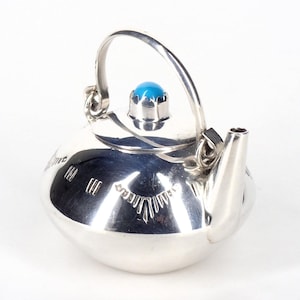 May include: A small, silver teapot with a rounded body and a curved handle. It features a turquoise stone on the lid and etched designs on the side. The spout is short and angled. The teapot is likely made of metal.