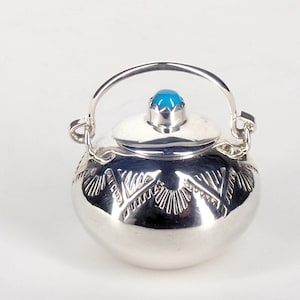 Op de afbeelding: Een kleine, ronde, zilverkleurige container met een deksel en handvat. Het deksel is bekroond met een turkooizen steen. De container heeft een decoratief patroon geëtst rond de basis. De handgreep is gebogen en aan de zijkanten bevestigd.